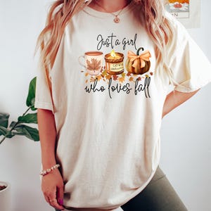 K&ouml;nnte beinhalten: Cremefarbenes T-Shirt mit dem Text "Just a girl who loves fall". Die Grafik zeigt eine Kaffeetasse, eine Kerze und einen K&uuml;rbis. Herbstliches Bekleidungsst&uuml;ck.