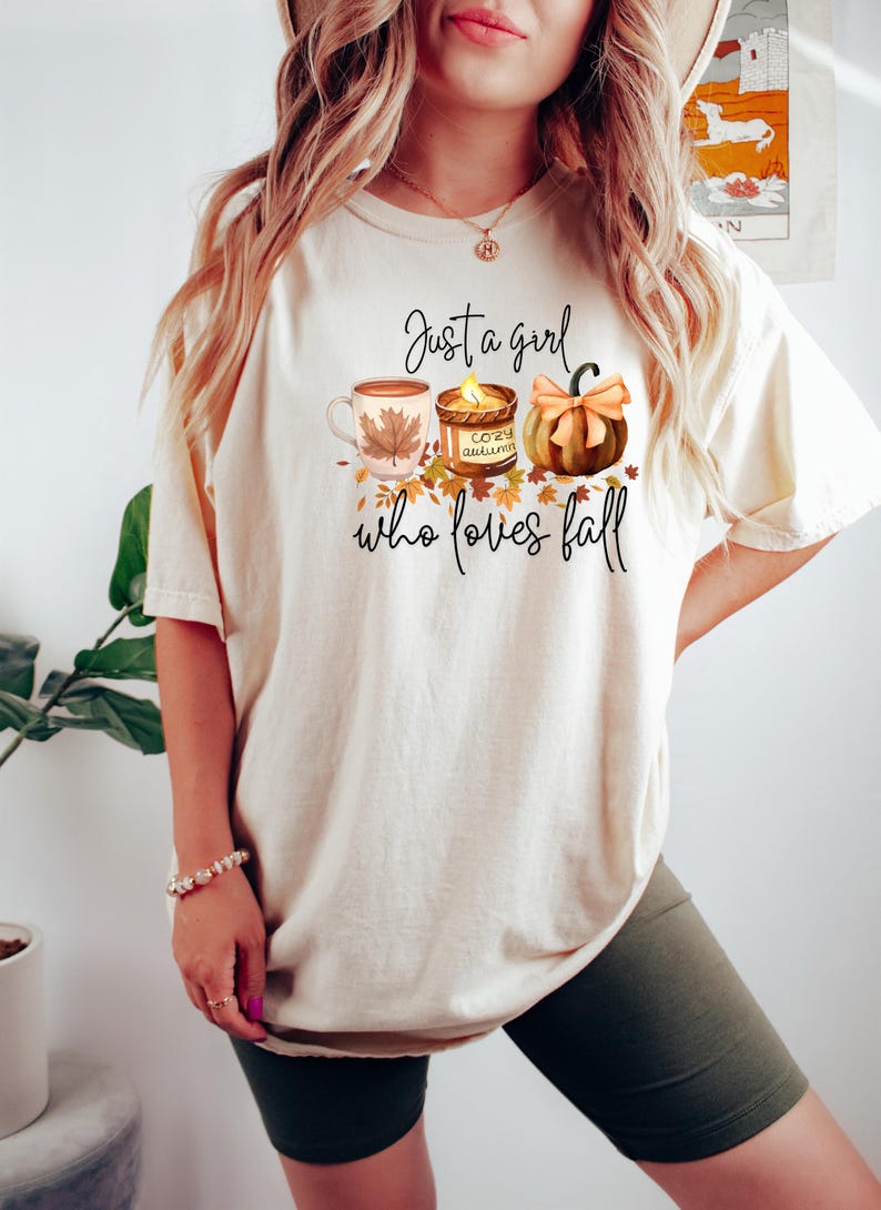 K&ouml;nnte beinhalten: Cremefarbenes T-Shirt mit dem Text "Just a girl who loves fall". Die Grafik zeigt eine Kaffeetasse, eine Kerze und einen K&uuml;rbis. Herbstliches Bekleidungsst&uuml;ck.
