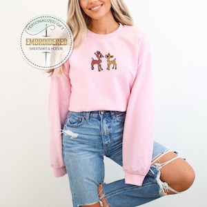 Peut inclure: Un sweat-shirt rose avec une image brod&eacute;e de deux cerfs de dessin anim&eacute;. Le sweat-shirt est port&eacute; par une personne aux cheveux longs blonds et un jean bleu.