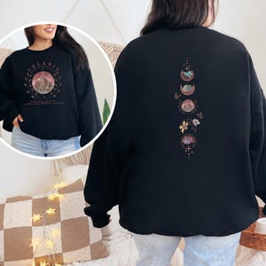 May include: Black sweatshirt with a circular graphic on the front featuring a mountain scene and the word "Velaris". The back of the sweatshirt has a vertical design of moon phases, flowers, and butterflies in shades of pink and gold.