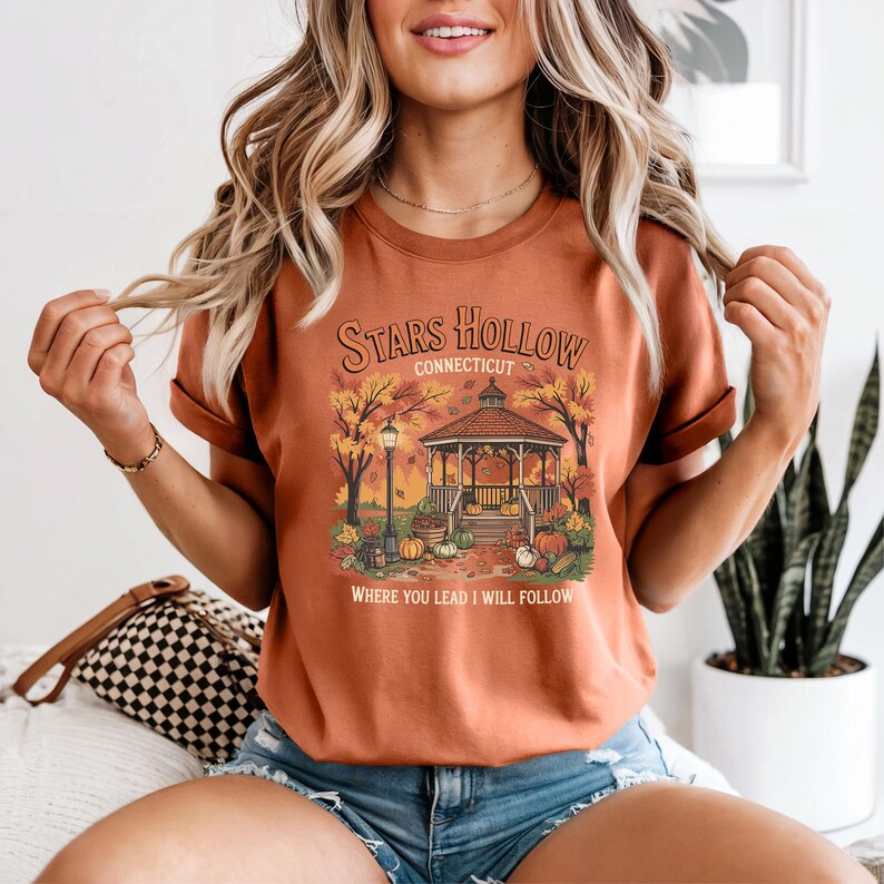 May include: Rust-colored t-shirt featuring a gazebo scene with the text "Stars Hollow Connecticut" and "Where You Lead I Will Follow." The design includes pumpkins, autumn leaves, and a lamp post, evoking a fall theme.