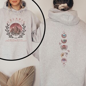 May include: Light gray hoodie with a front graphic of a mountain scene within a circle, the text "VELARIS CITY OF STARLIGHT" and a quote. The back features a vertical design of moon phases with nature-themed illustrations.