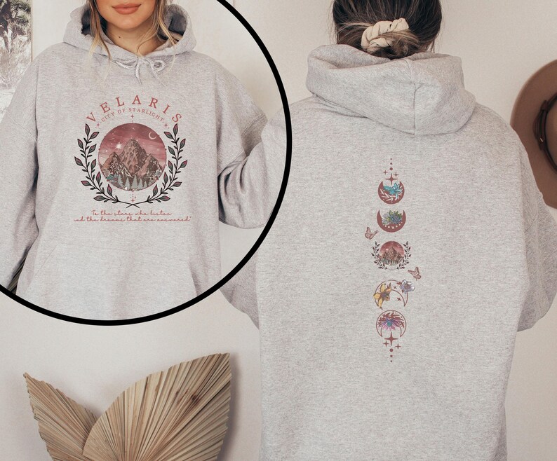May include: Light gray hoodie with a front graphic of a mountain scene within a circle, the text "VELARIS CITY OF STARLIGHT" and a quote. The back features a vertical design of moon phases with nature-themed illustrations.