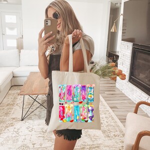 Pu&ograve; includere: Una borsa tote color crema con illustrazioni colorate di bevande tropicali. La borsa ha una forma rettangolare con due manici lunghi. Le bevande sono di vari colori, con accenti di frutta e fiori. La borsa &egrave; tenuta da una persona.