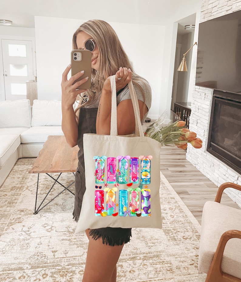 Pu&ograve; includere: Una borsa tote color crema con illustrazioni colorate di bevande tropicali. La borsa ha una forma rettangolare con due manici lunghi. Le bevande sono di vari colori, con accenti di frutta e fiori. La borsa &egrave; tenuta da una persona.