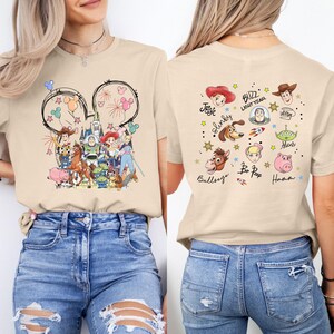 Puede incluir: Camiseta beige con un gr&aacute;fico colorido de los personajes de Toy Story de Disney en la parte delantera y trasera. El dise&ntilde;o frontal incluye orejas de Mickey Mouse con fuegos artificiales e ilustraciones de personajes. La parte posterior muestra nombres e ilustraciones de personajes.