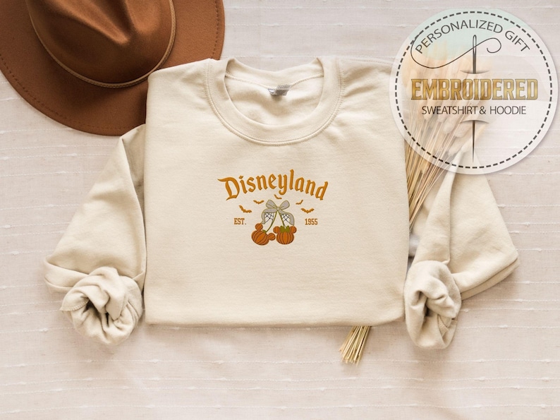 Pode incluir: Camisola creme com bordado laranja "Disneyland", adornada com ab&oacute;boras e morcegos. A parte superior de manga comprida tem gola redonda. Um chap&eacute;u castanho &eacute; vis&iacute;vel no fundo. O design inclui o ano de 1955.
