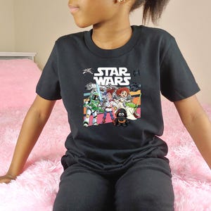 Puede incluir: Camiseta negra de Star Wars con un gr&aacute;fico colorido de personajes de la pel&iacute;cula. La camiseta tiene el logo de Star Wars en blanco. La persona lleva pantalones negros y est&aacute; sentada en una manta rosa y esponjosa.