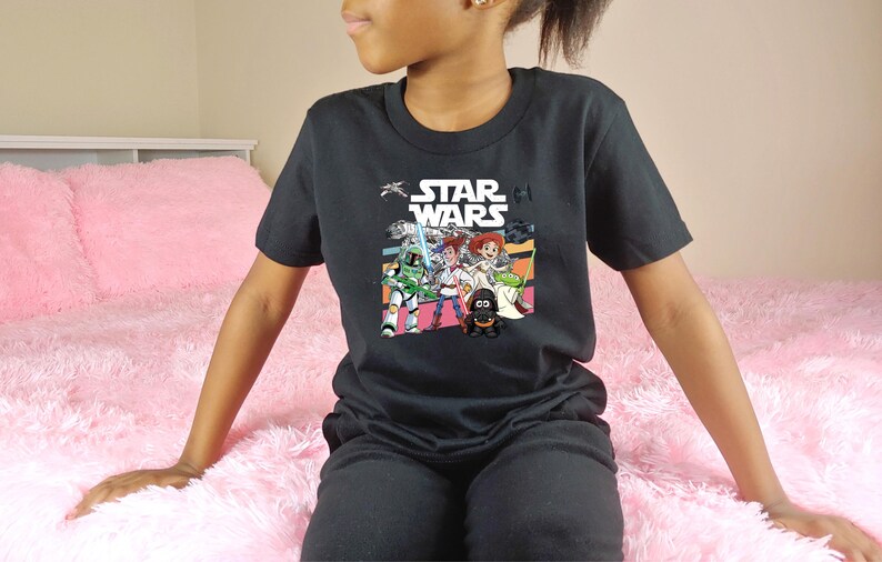 Puede incluir: Camiseta negra de Star Wars con un gr&aacute;fico colorido de personajes de la pel&iacute;cula. La camiseta tiene el logo de Star Wars en blanco. La persona lleva pantalones negros y est&aacute; sentada en una manta rosa y esponjosa.