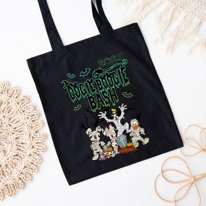 May include: Black tote bag with the text "2025 Oogie Boogie Bash" in green. The bag features cartoon characters dressed as mummies and other Halloween costumes. The bag has black handles.