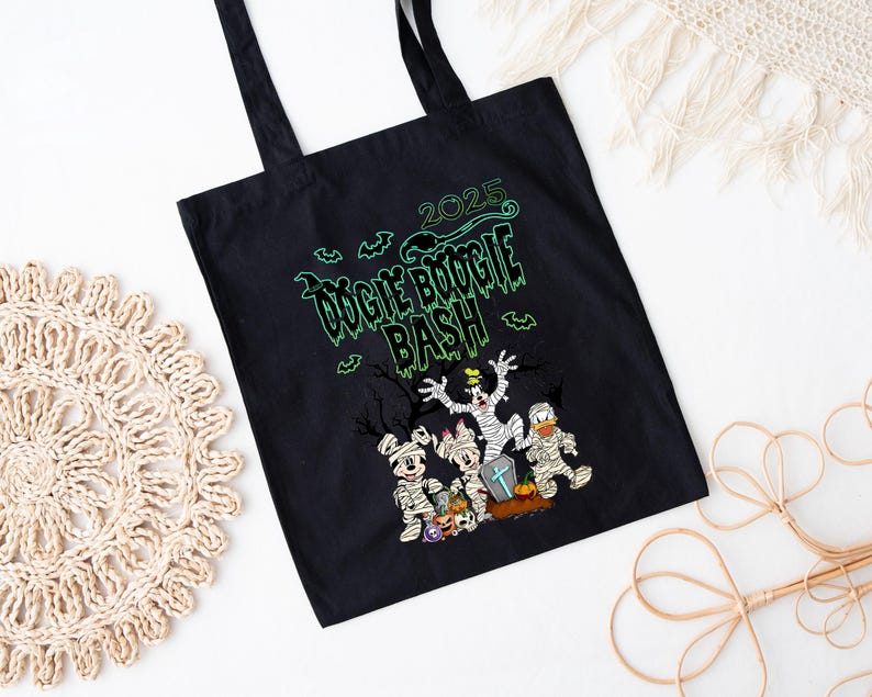 May include: Black tote bag with the text "2025 Oogie Boogie Bash" in green. The bag features cartoon characters dressed as mummies and other Halloween costumes. The bag has black handles.