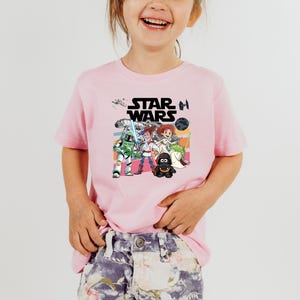 Puede incluir: Camiseta rosa de manga corta con un gr&aacute;fico colorido de personajes de Star Wars. El dise&ntilde;o incluye el logotipo de Star Wars y varias figuras de dibujos animados de la franquicia. La camiseta la lleva un ni&ntilde;o.