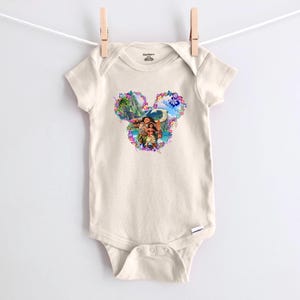 Puede incluir: Un body de beb&eacute; color crema con un gr&aacute;fico colorido de tem&aacute;tica Disney. El gr&aacute;fico presenta una forma de cabeza de Mickey Mouse floral con im&aacute;genes de personajes y paisajes tropicales. El body est&aacute; colgado de un tendedero con una pinza de madera.