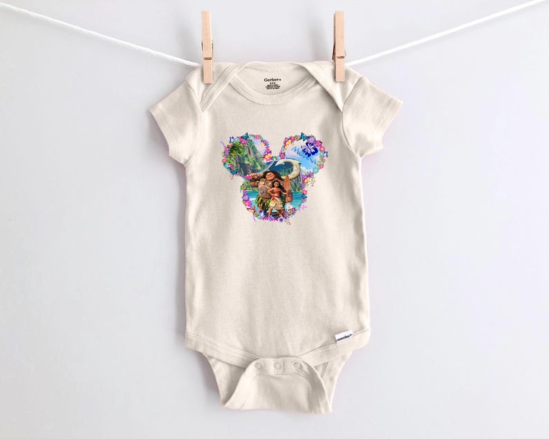 Puede incluir: Un body de beb&eacute; color crema con un gr&aacute;fico colorido de tem&aacute;tica Disney. El gr&aacute;fico presenta una forma de cabeza de Mickey Mouse floral con im&aacute;genes de personajes y paisajes tropicales. El body est&aacute; colgado de un tendedero con una pinza de madera.