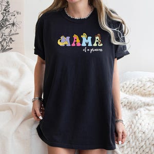 K&ouml;nnte beinhalten: Schwarzes T-Shirt mit einer bunten Grafik, auf der "Mama of a princess" mit Disney-Prinzessinnen-Charakteren steht.