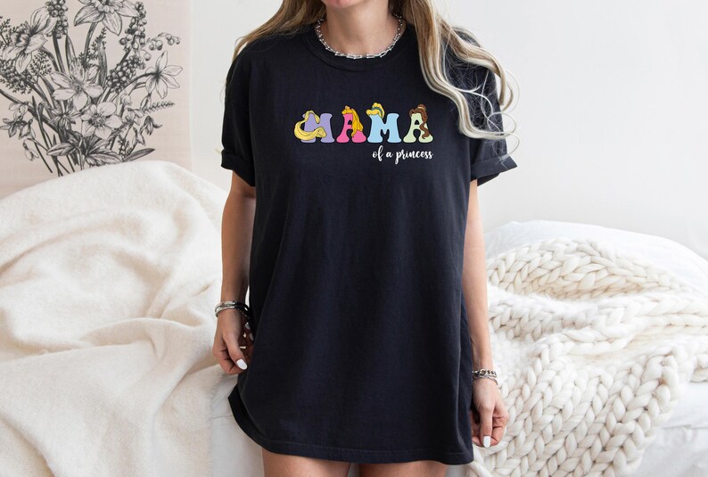 K&ouml;nnte beinhalten: Schwarzes T-Shirt mit einer bunten Grafik, auf der "Mama of a princess" mit Disney-Prinzessinnen-Charakteren steht.