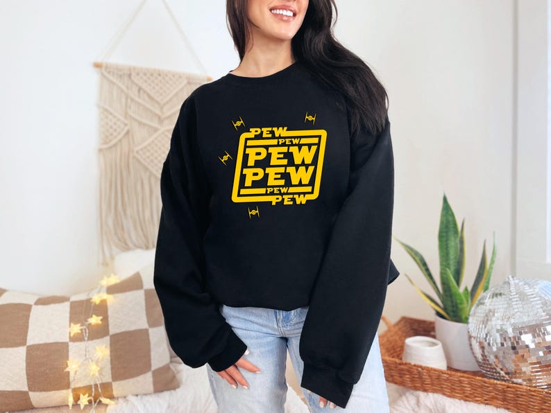 May include: Black sweatshirt with a yellow graphic that says "PEW PEW PEW" in a square. The design includes small yellow starfighter illustrations. The sweatshirt is a crew neck style.