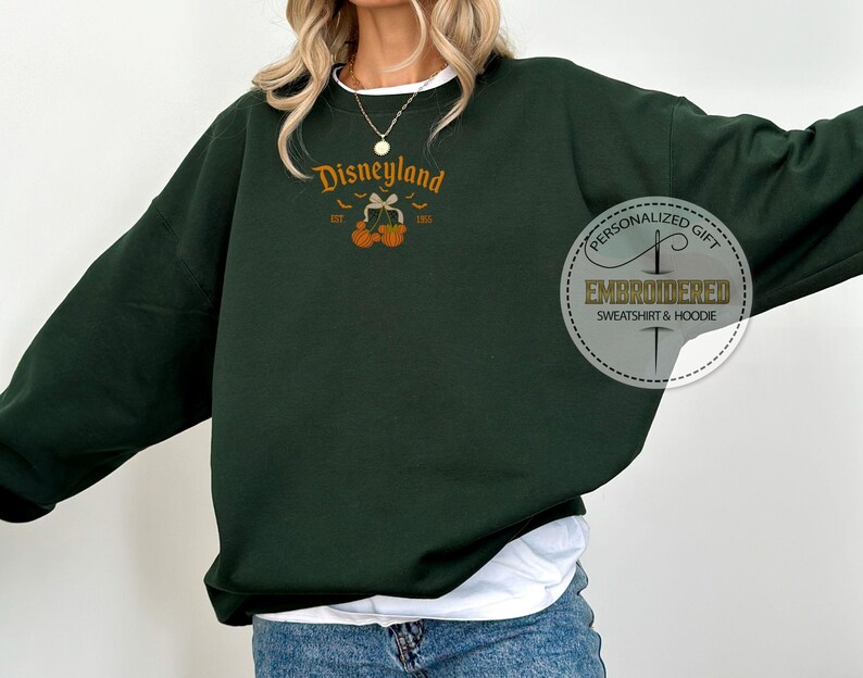 Pode incluir: Camisola verde escuro com a palavra "Disneyland" em escrita laranja, um design de la&ccedil;o e ab&oacute;boras, e o texto "EST 1955". Um autocolante circular &agrave; direita diz "PERSONALIZED GIFT EMBROIDERED SWEATSHIRT & HOODIE".