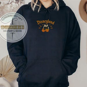 Pode incluir: Um moletom azul marinho com um design da Disneyland. O design inclui a palavra "Disneyland" em letras douradas, com morcegos e ab&oacute;boras. O moletom tamb&eacute;m tem o texto "EST 1955". A imagem tamb&eacute;m inclui um logotipo de presente personalizado.