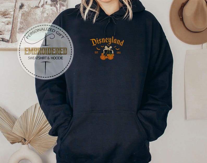Pode incluir: Um moletom azul marinho com um design da Disneyland. O design inclui a palavra "Disneyland" em letras douradas, com morcegos e ab&oacute;boras. O moletom tamb&eacute;m tem o texto "EST 1955". A imagem tamb&eacute;m inclui um logotipo de presente personalizado.