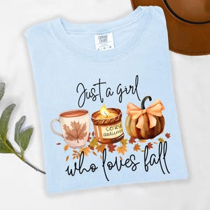 K&ouml;nnte beinhalten: Hellblaues T-Shirt mit dem Text "Just a girl who loves fall". Die Grafik zeigt eine Kaffeetasse, eine Kerze und einen K&uuml;rbis in herbstlichen Farben.