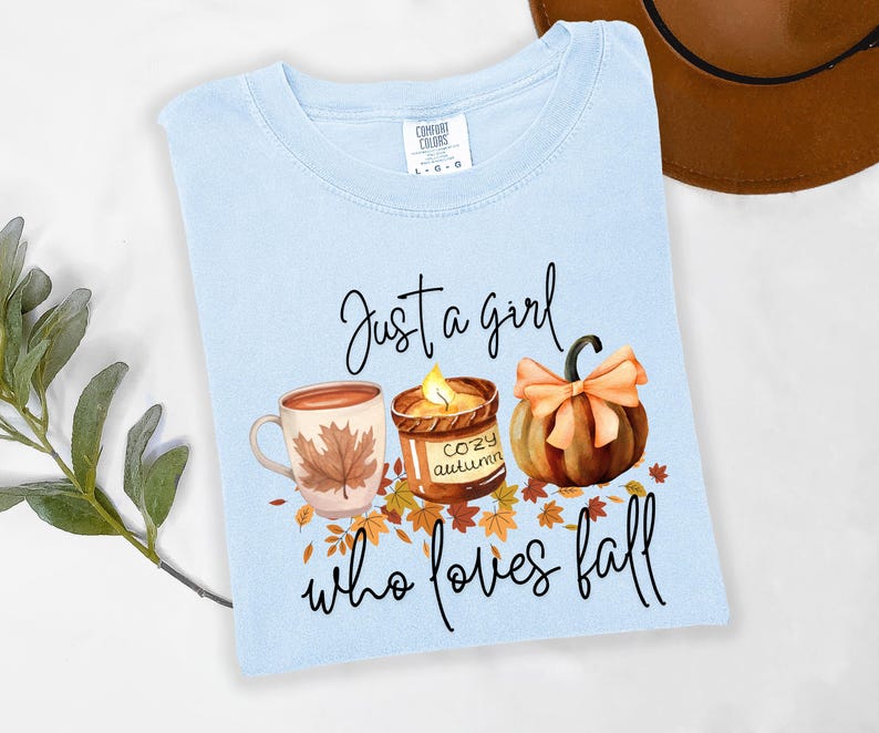 K&ouml;nnte beinhalten: Hellblaues T-Shirt mit dem Text "Just a girl who loves fall". Die Grafik zeigt eine Kaffeetasse, eine Kerze und einen K&uuml;rbis in herbstlichen Farben.