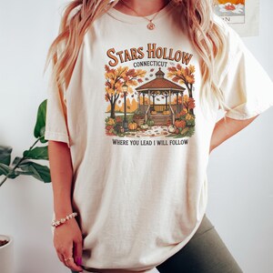 May include: Cream-colored t-shirt featuring a fall-themed graphic with a gazebo, pumpkins, and autumn foliage. The text reads "Stars Hollow Connecticut" and "Where You Lead I Will Follow."