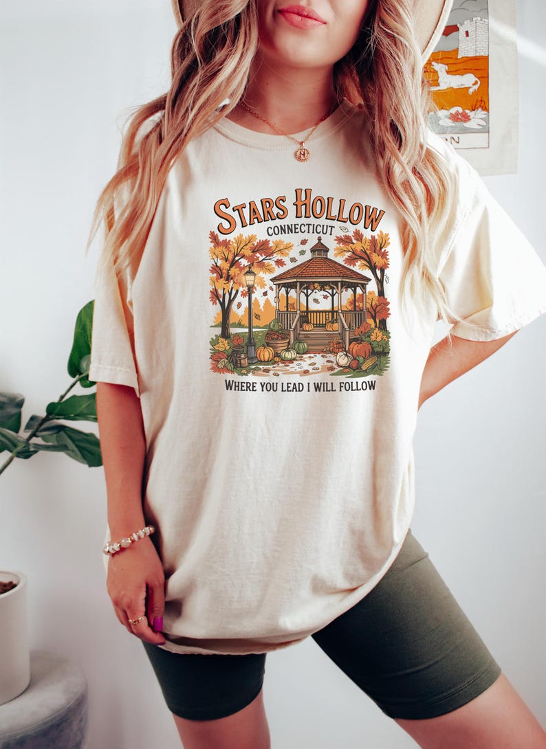 May include: Cream-colored t-shirt featuring a fall-themed graphic with a gazebo, pumpkins, and autumn foliage. The text reads "Stars Hollow Connecticut" and "Where You Lead I Will Follow."
