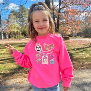 May include: A bright pink Star Wars sweatshirt featuring various character graphics, including a Stormtrooper, Ewoks, and Darth Vader's helmet. The sweatshirt has long sleeves and a crew neck. The words "STAR WARS" are printed on the front.