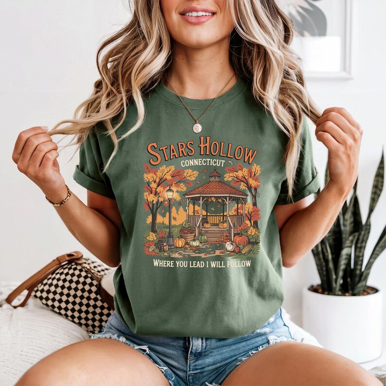 May include: Green t-shirt with a graphic featuring a gazebo, pumpkins, and fall foliage. The text reads "Stars Hollow Connecticut" and "Where You Lead I Will Follow". The shirt has rolled-up sleeves.