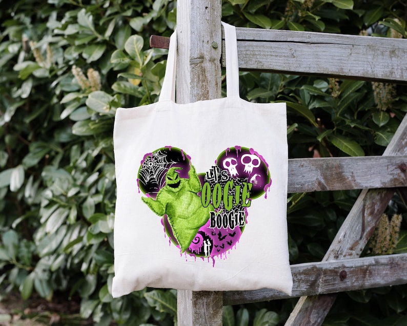 May include: A white tote bag with a green and black Oogie Boogie design and the text "Let's Oogie Boogie". The design includes a spiderweb, skulls, and a purple dripping effect. The bag is hanging on a wooden fence.