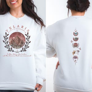 May include: White sweatshirt with a front graphic featuring the text "VELARIS" and a mountain scene within a circular design. The back of the sweatshirt displays a vertical design of moon phases. The text "In the stars who listen and the dreams that are answered" is also present.