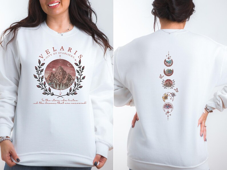 May include: White sweatshirt with a front graphic featuring the text "VELARIS" and a mountain scene within a circular design. The back of the sweatshirt displays a vertical design of moon phases. The text "In the stars who listen and the dreams that are answered" is also present.