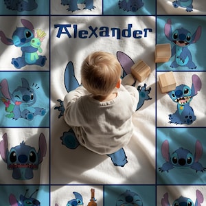 May include: A baby blanket featuring a blue cartoon character design and the name "Alexander" in blue text. The blanket has a white background with blue squares and cartoon character illustrations. Wooden blocks are also present.