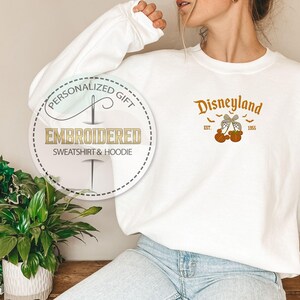 Pode incluir: Camisola branca bordada com a palavra "Disneyland" em laranja, juntamente com ab&oacute;boras e morcegos. A camisola tamb&eacute;m tem um gr&aacute;fico circular que diz "Presente Personalizado" e "Camisola e Moletom Bordados".