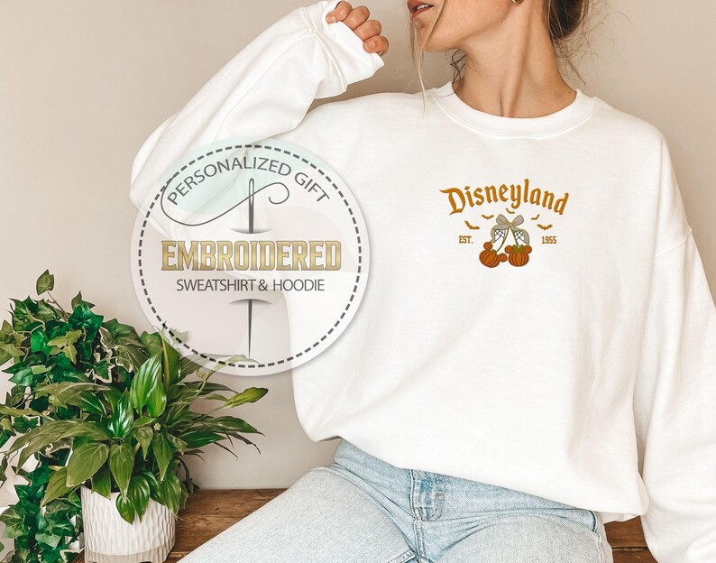 Pode incluir: Camisola branca bordada com a palavra "Disneyland" em laranja, juntamente com ab&oacute;boras e morcegos. A camisola tamb&eacute;m tem um gr&aacute;fico circular que diz "Presente Personalizado" e "Camisola e Moletom Bordados".