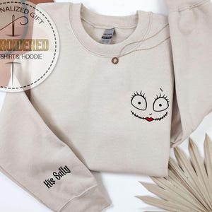 May include: Beige sweatshirt with a Sally face design embroidered on the front. The design features large eyes, a stitched mouth, and red lips. The sleeve reads "His Sally" in black script. A gold necklace is draped on the sweatshirt.