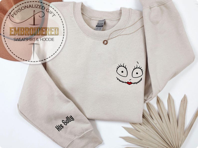 May include: Beige sweatshirt with a Sally face design embroidered on the front. The design features large eyes, a stitched mouth, and red lips. The sleeve reads "His Sally" in black script. A gold necklace is draped on the sweatshirt.