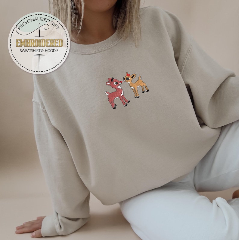 Peut inclure: Un sweat-shirt beige avec un motif brod&eacute; de deux rennes qui s'embrassent. Les rennes sont bruns et rouges avec des accents blancs. Le sweat-shirt a un col rond et des manches longues.