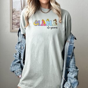 K&ouml;nnte beinhalten: Ein hellgr&uuml;nes T-Shirt mit einer Grafik des Wortes "MAMA", das mit Disney-Prinzessinnen ausgeschrieben ist. Der Text "of a princess" steht unter der Grafik.