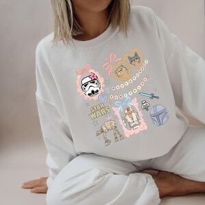 May include: White sweatshirt featuring Star Wars characters and elements in pastel colors. The design includes a Stormtrooper helmet, R2-D2, an AT-AT, and other graphics within decorative frames. The words "STAR WARS" are also included.