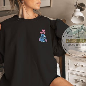 May include: Black sweatshirt featuring an embroidered Stitch character with pink hearts. The design is on the upper left chest. A circular graphic on the right side reads "Personalized Gift, Embroidered Sweatshirt & Hoodie."