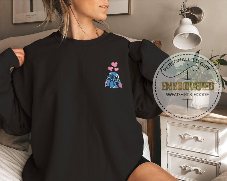 May include: Black sweatshirt featuring an embroidered Stitch character with pink hearts. The design is on the upper left chest. A circular graphic on the right side reads "Personalized Gift, Embroidered Sweatshirt & Hoodie."