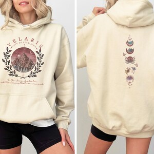 May include: Cream-colored hoodie with a front graphic featuring a mountain scene within a wreath and the text "VELARIS CITY OF STARLIGHT". The back displays a vertical design of moon phases and floral elements. The hoodie has a front pocket and a hood.