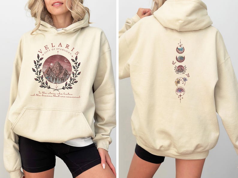 May include: Cream-colored hoodie with a front graphic featuring a mountain scene within a wreath and the text "VELARIS CITY OF STARLIGHT". The back displays a vertical design of moon phases and floral elements. The hoodie has a front pocket and a hood.