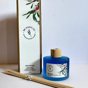 Peut inclure: Un flacon diffuseur de roseaux en verre bleu avec un couvercle en bois et une étiquette indiquant "BEL SCENTS CO. HANDMADE ARTISAN CANDLES GUM LEAVES Reed Diffuser Blends of Lemon Myrtle, Eucalyptus & Blue Cypress Pine 200ml". Le flacon est posé sur une surface blanche avec un jeu de bâtonnets de roseaux en bois.