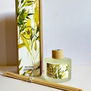 Könnte beinhalten: Eine klare Glasflasche mit Schilfrohrdiffusor mit einem braunen Holzdeckel und einem Etikett mit der Aufschrift "Lemongrass & Lime". Die Flasche steht auf einer weißen Oberfläche mit einer Schachtel dahinter. Die Schachtel ist braun mit einem Blumendesign und dem Text "Bel Scents Co. Handmade Artisan Candles".