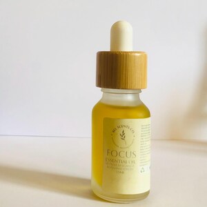 May include: A small, frosted glass bottle of essential oil with a wooden cap and dropper. The bottle contains a yellow liquid and has a label that reads "FOCUS Essential Oil". The label also includes the brand name "Bee Scents Co."