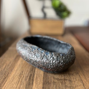 May include: A small, oval-shaped ceramic planter with a rough, textured surface. The planter is dark grey with hints of blue and red.