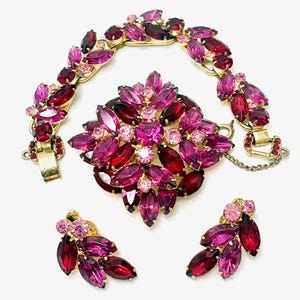 May include: A jewelry set consisting of a gold-tone bracelet, a large brooch, and a pair of clip-on earrings, all featuring marquise-cut and round-cut crystals in various shades of pink, magenta, and deep red, set in a floral-inspired arrangement.